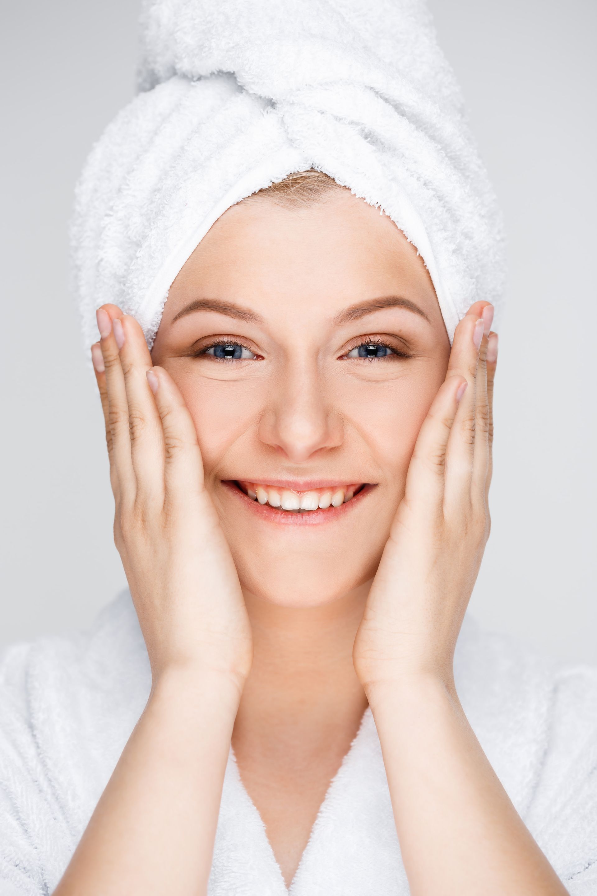 A woman with a towel wrapped around her head is smiling and touching her face.