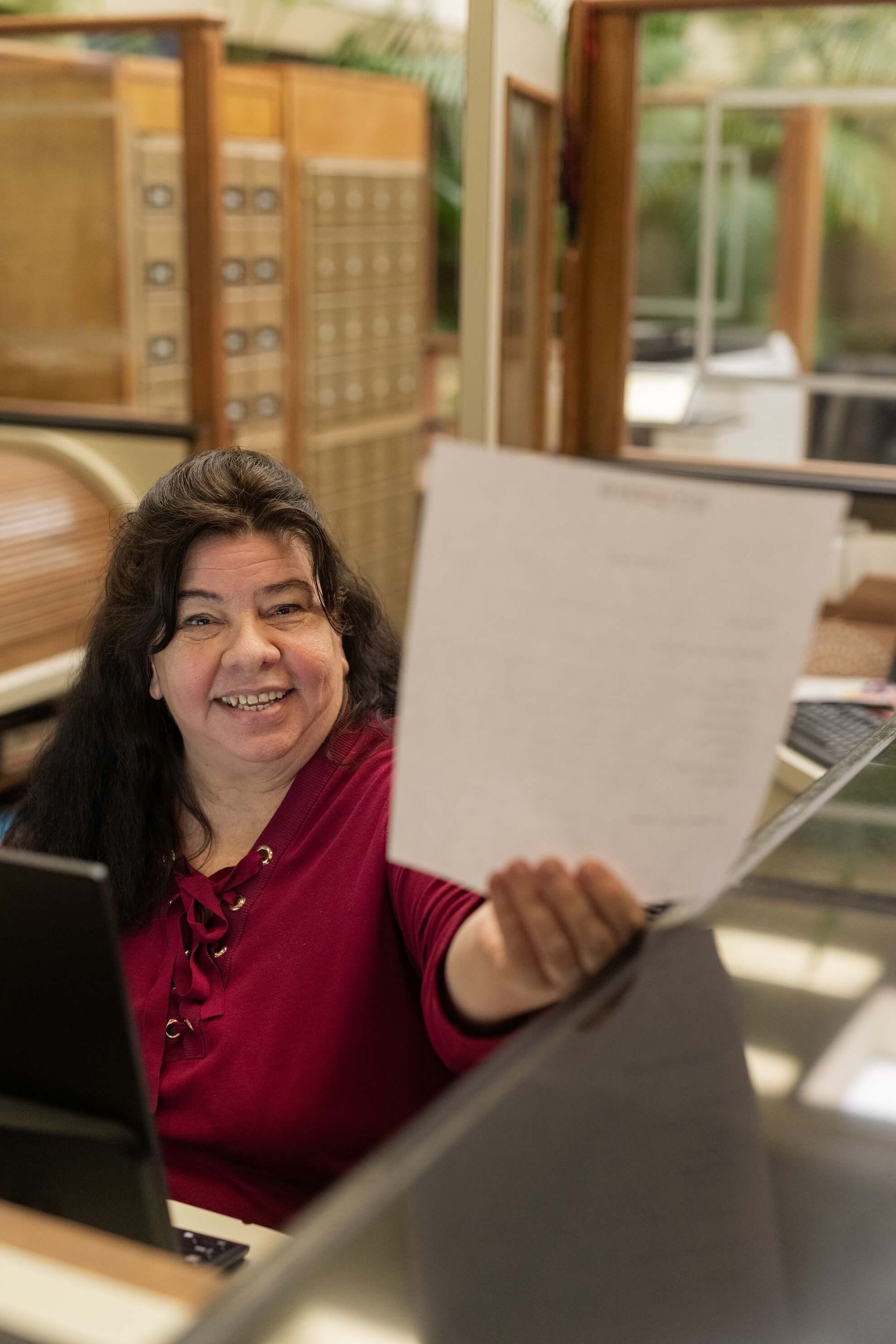 Woman in suit wearing headset smiles while working at a computer in an office setting.