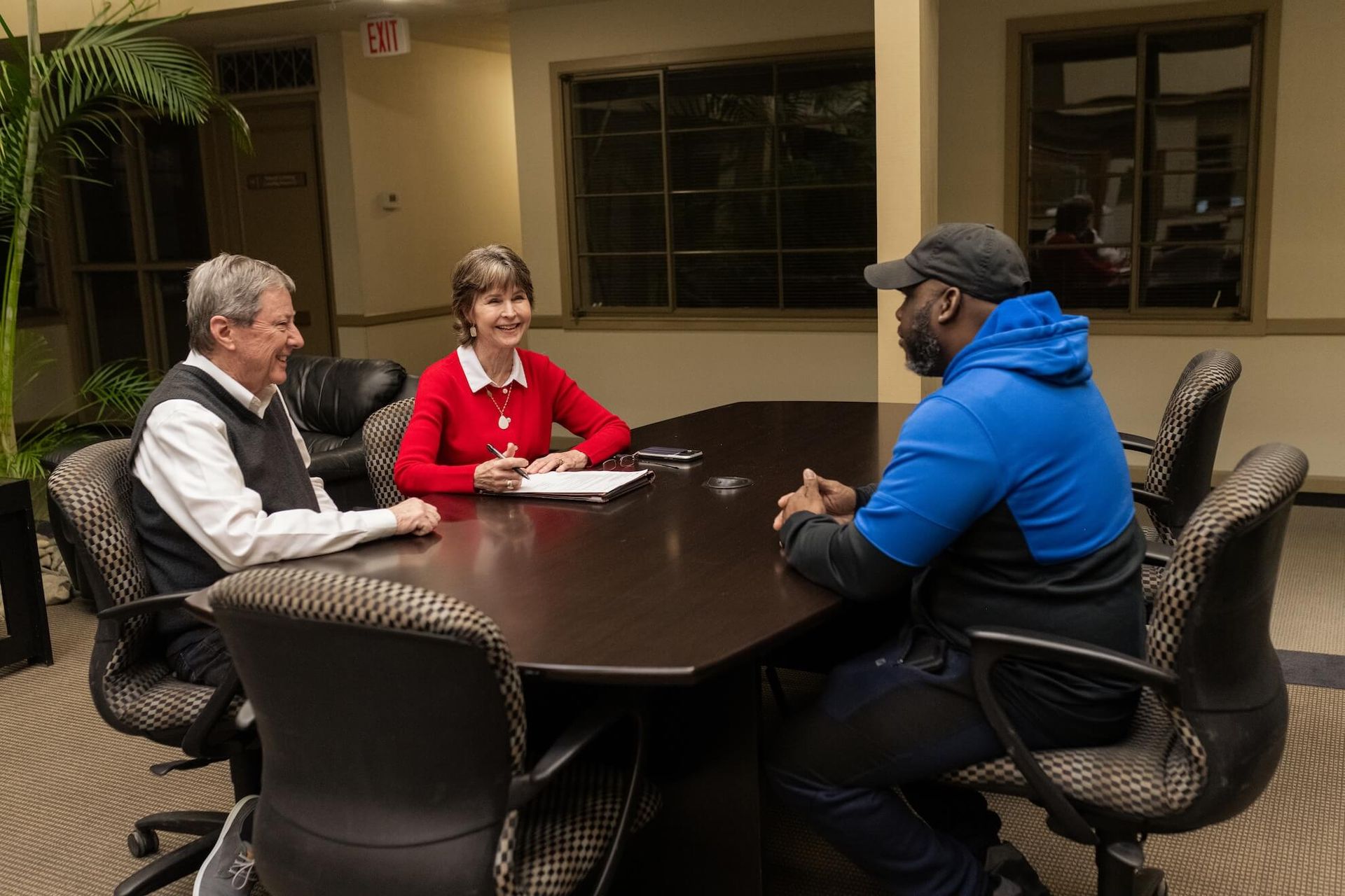 Three people sit around a table in an office, engaged in a discussion while looking at documents.