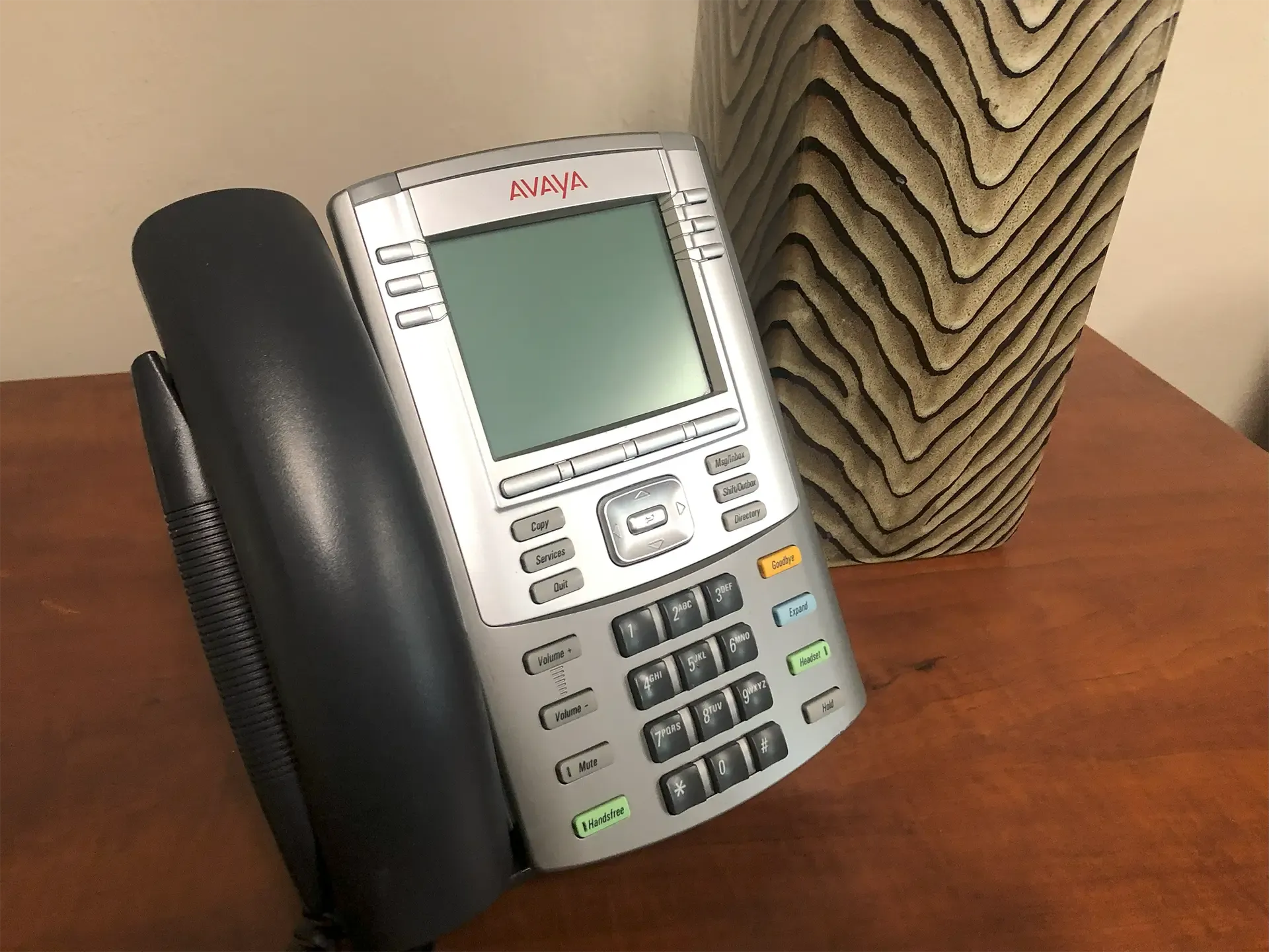 Gray and silver Avaya desk phone on a wooden surface next to a decorative vase.