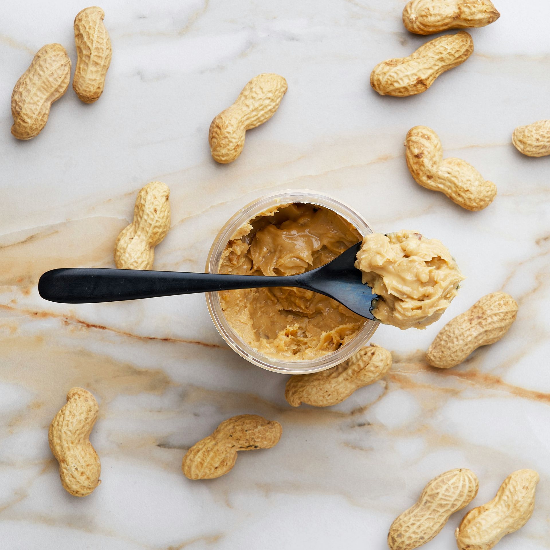 Peanut butter in a bowl with a spoon, surrounded by scattered peanuts on a marble surface