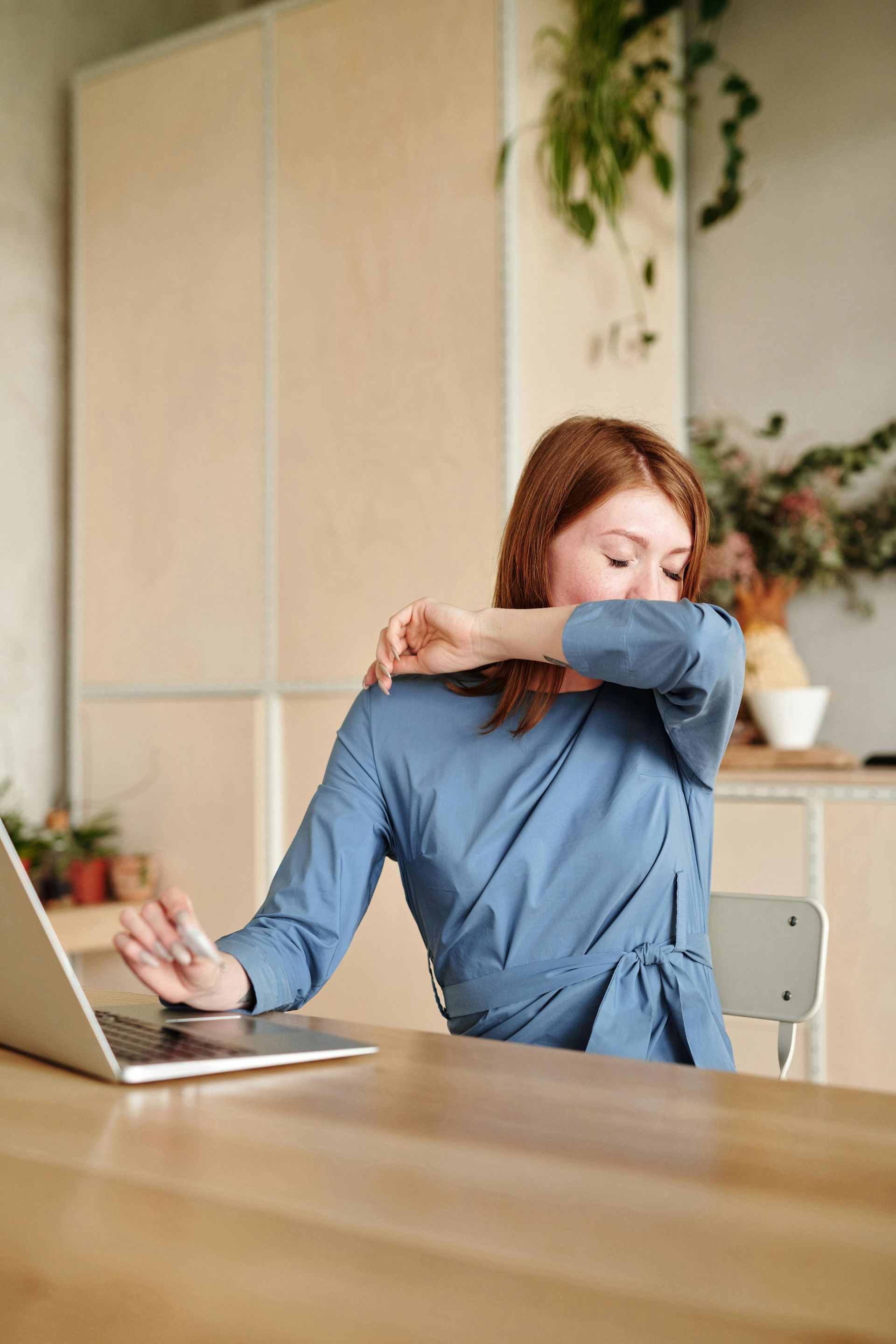 Woman at a desk sneezing into her elbow beside an open laptop in a bright office