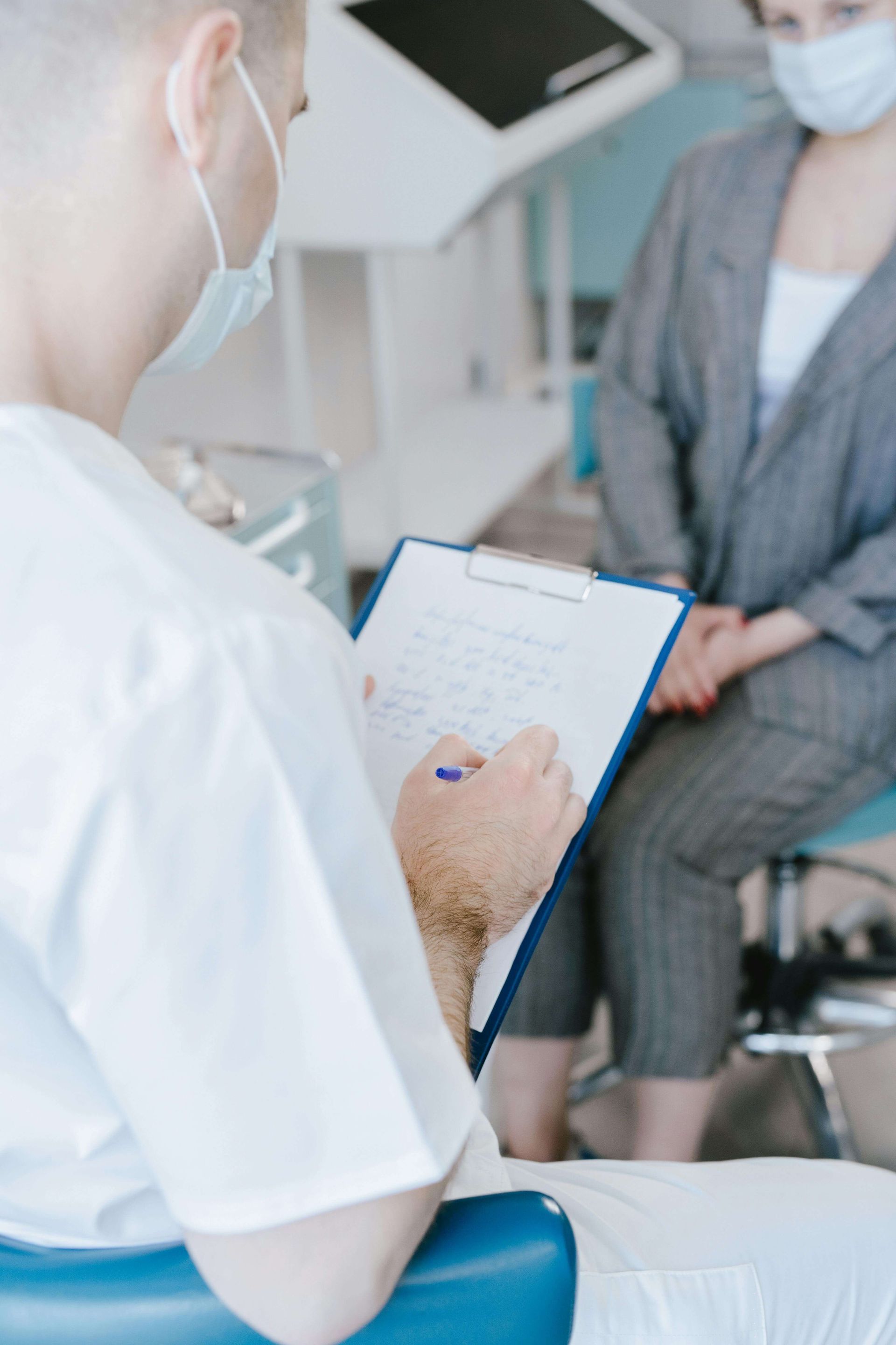Doctor taking notes during a consultation with a masked patient in a clinic.