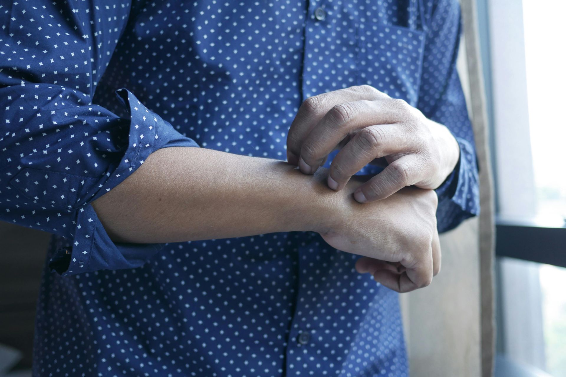 Person in a blue polka-dot shirt clutching their forearm near a window
