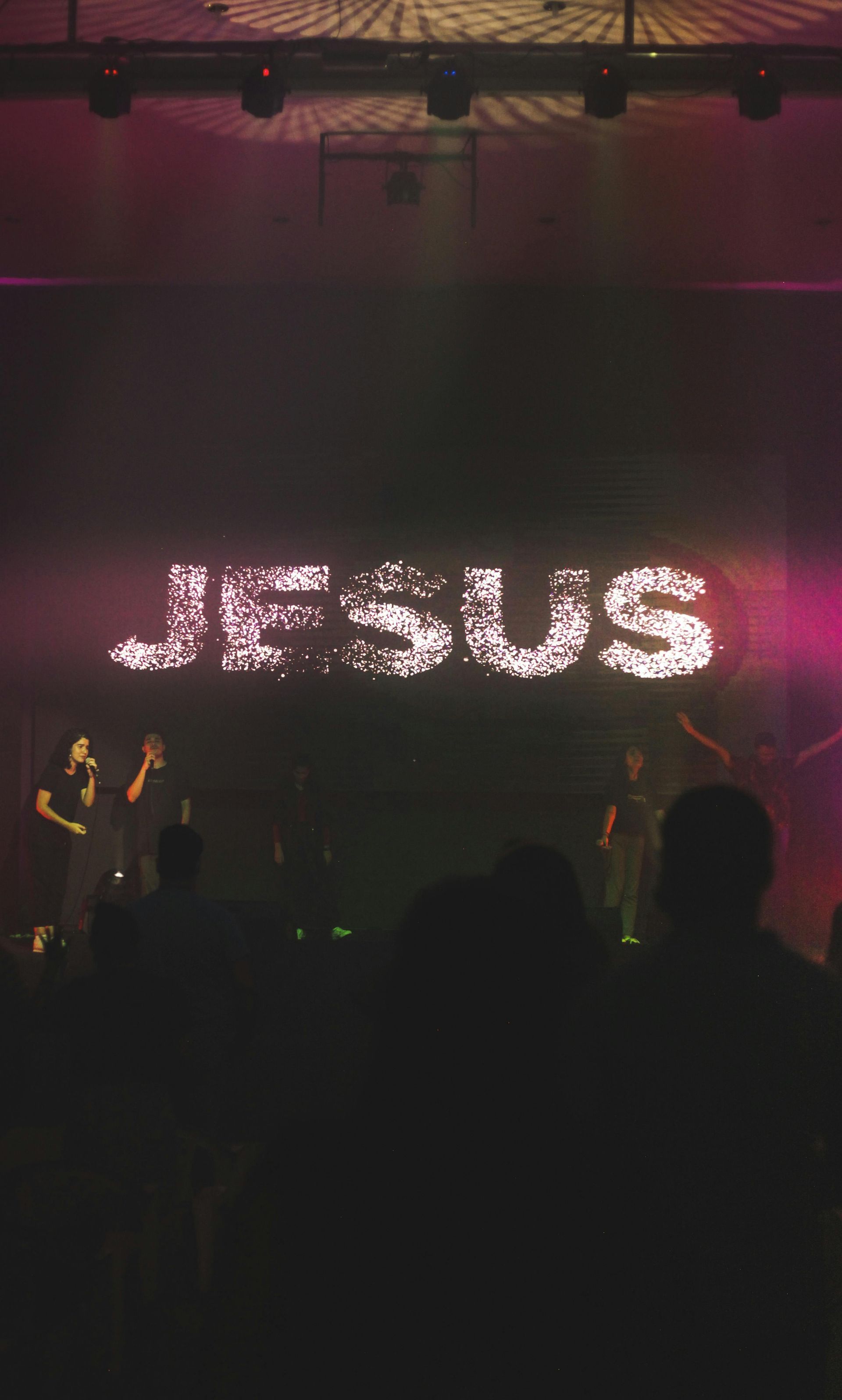 Concert stage with bright pink “JESUS” sign and silhouetted performers in the foreground