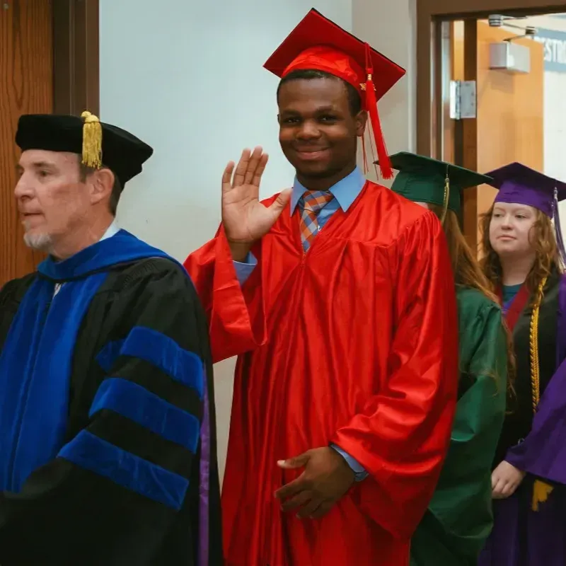Graduates in caps and gowns waving in a hallway, led by a faculty member in academic regalia