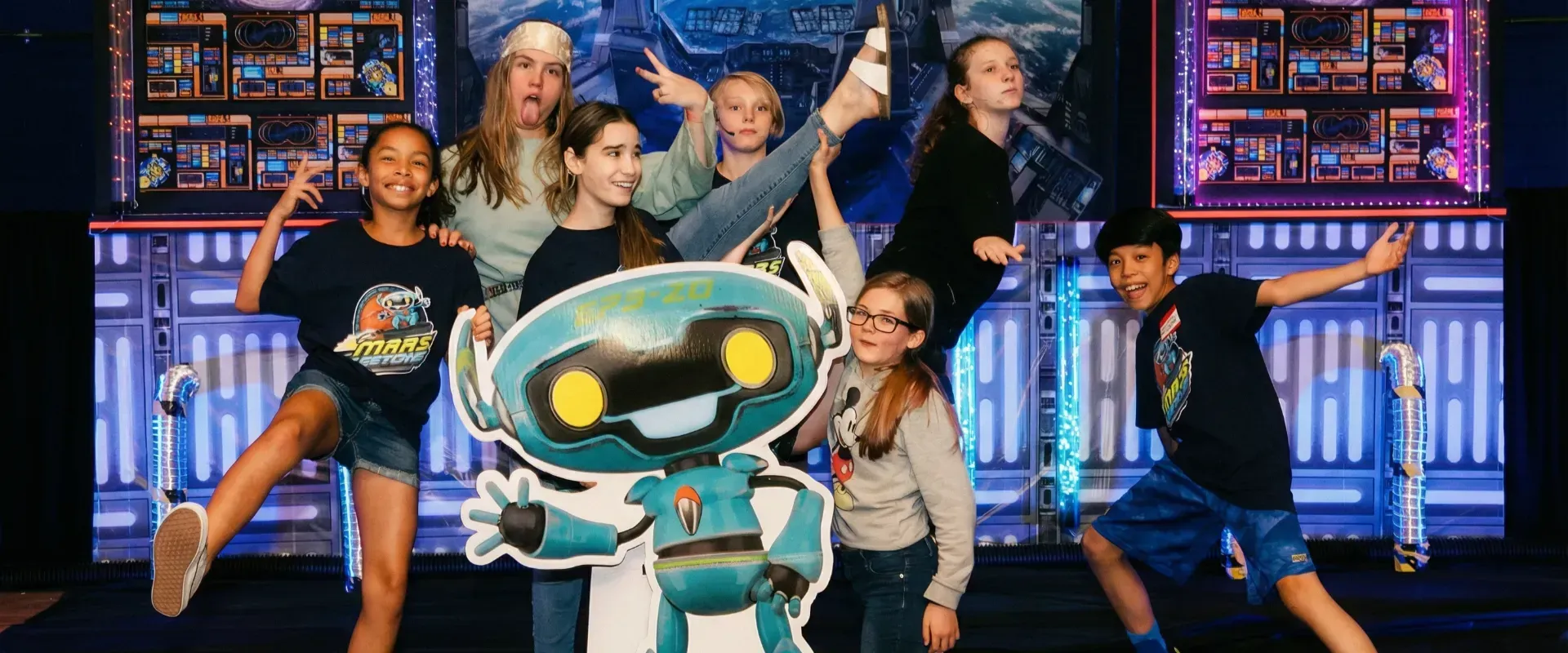 Cartoon robot in front of a colorful group pose on a glowing stage set