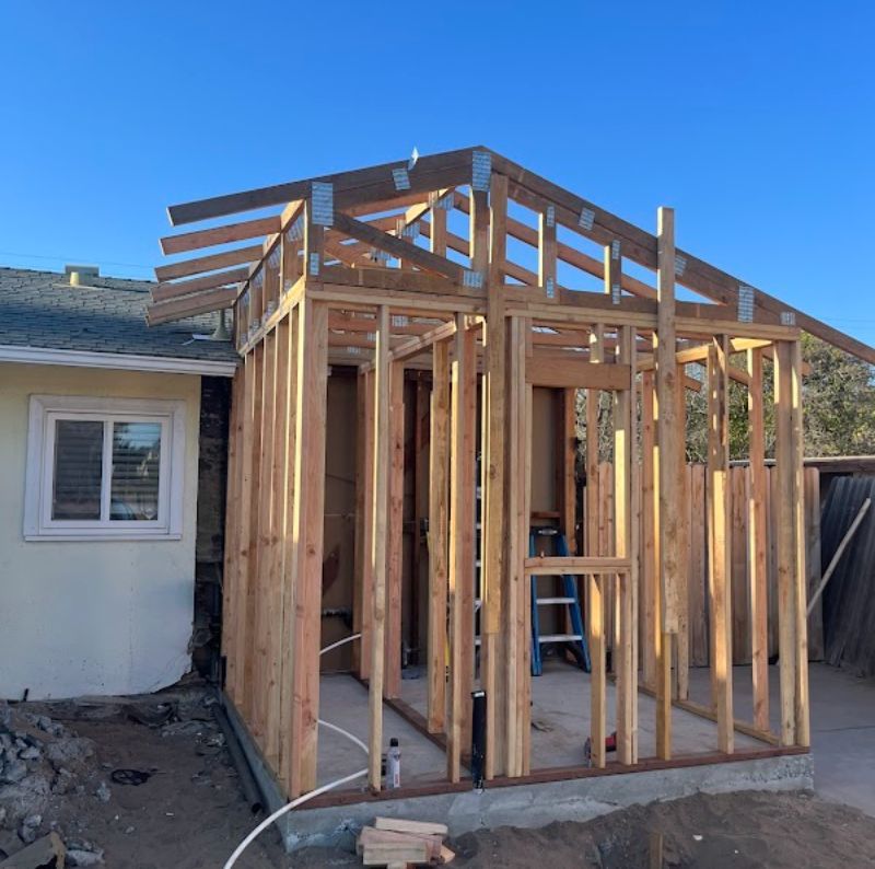 Framed wooden home addition under construction beside a beige house under a clear blue sky
