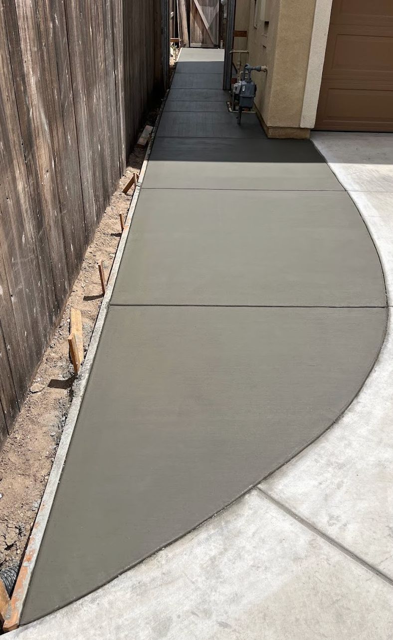 Freshly poured gray concrete sidewalk beside a wooden fence and building wall in a narrow passage