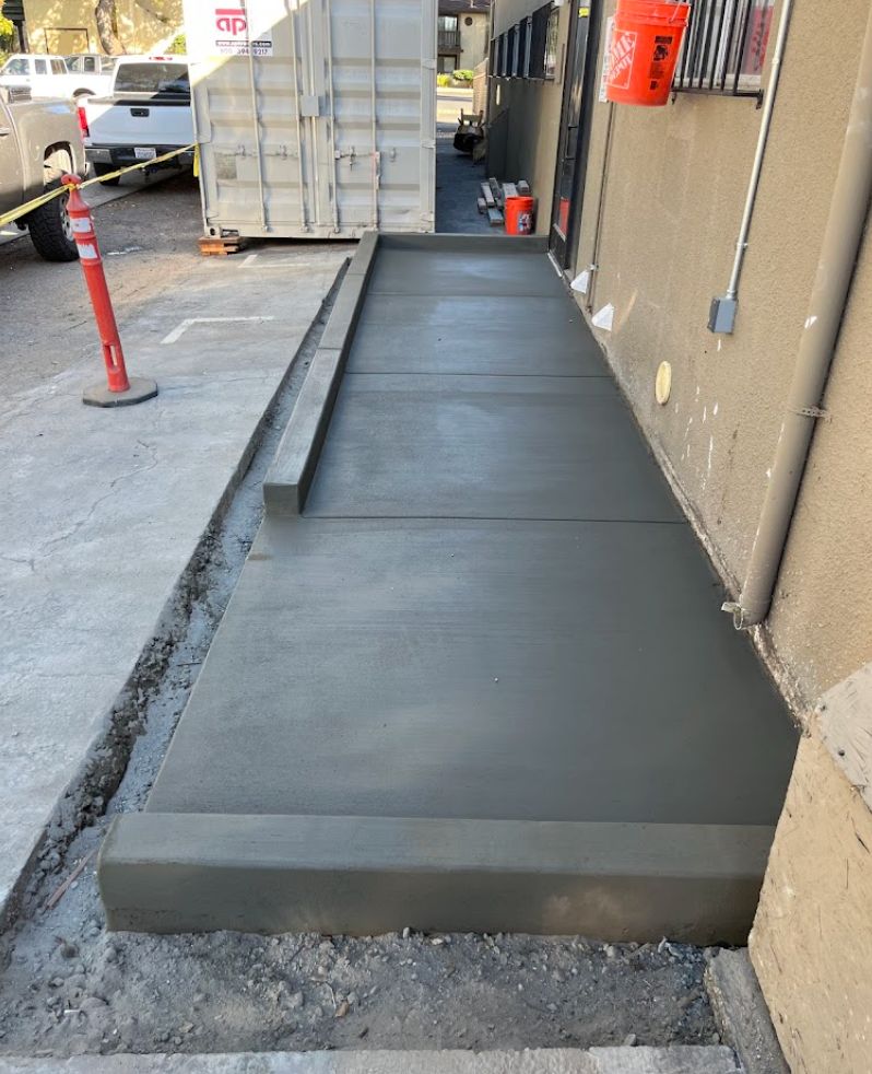 Freshly poured concrete walkway beside a building under construction, next to a curb and utility poles