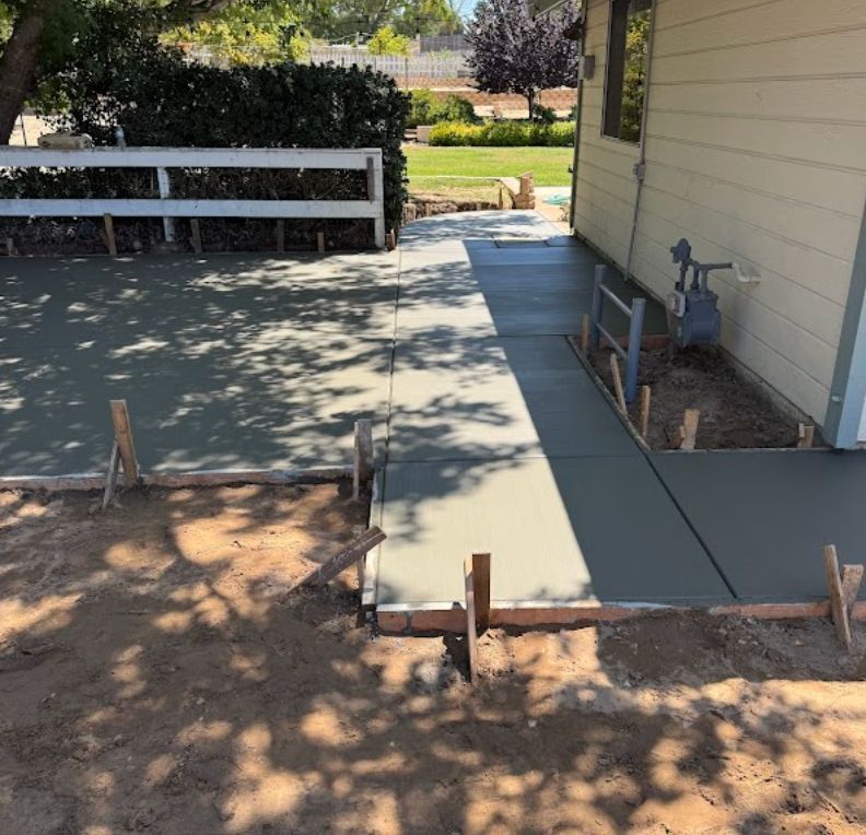 Fresh gray concrete patio walkway beside a house, with forms and a bench in a shaded backyard.