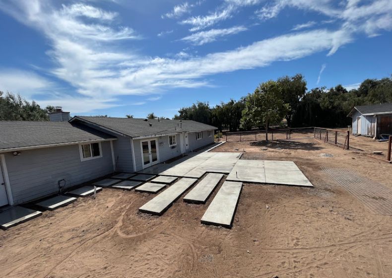 Single-story house with damaged roof panels lying in the dirt yard under a blue sky