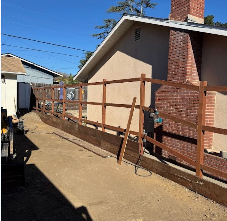 Wooden handrail along a dirt path beside a house and brick chimney under a clear blue sky