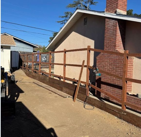 Wooden handrail along a dirt path beside a house and brick chimney under a clear blue sky