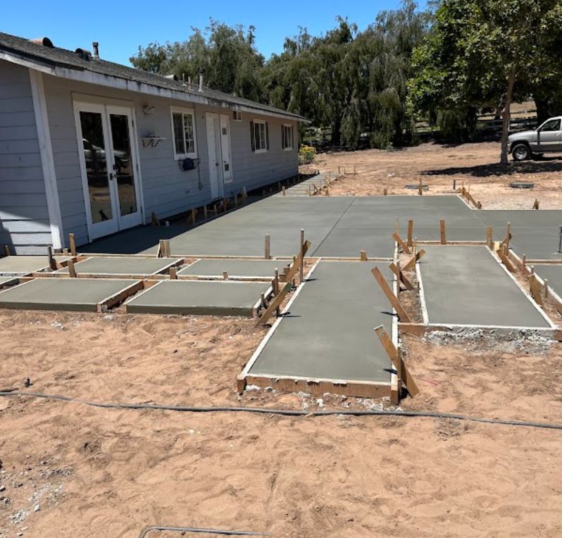Fresh concrete walkway and pads under construction beside a white house on a dirt lot