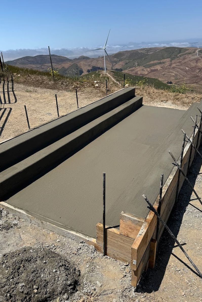 Fresh concrete foundation under construction with rebar on a hillside overlooking mountains and wind turbines
