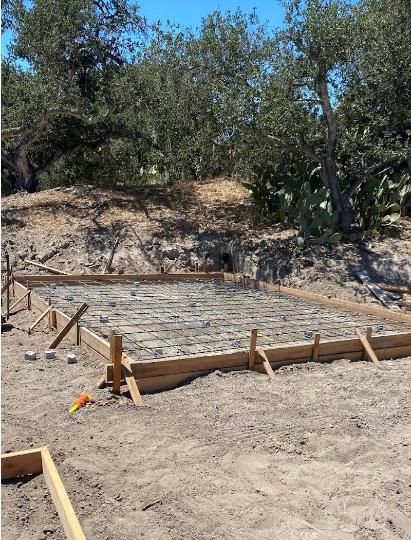Wooden formwork for a concrete slab on a dirt lot with trees and brush in the background
