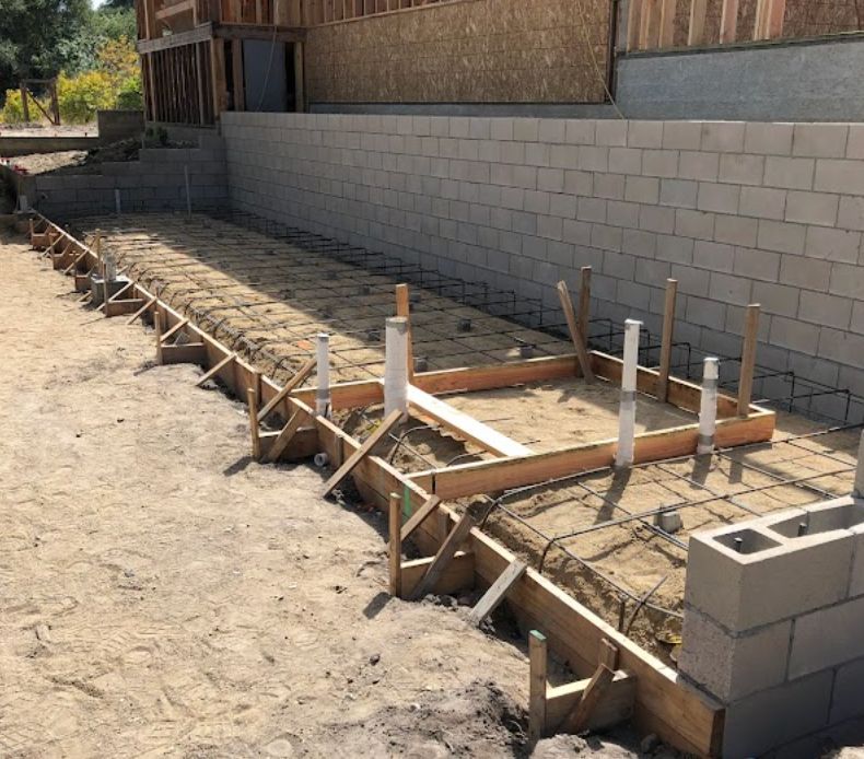 Concrete foundation formwork and rebar set beside a cinder block wall in a dusty yard