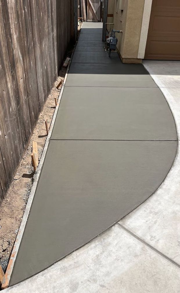 Freshly poured concrete walkway beside a wooden fence and building wall with a curved edge.