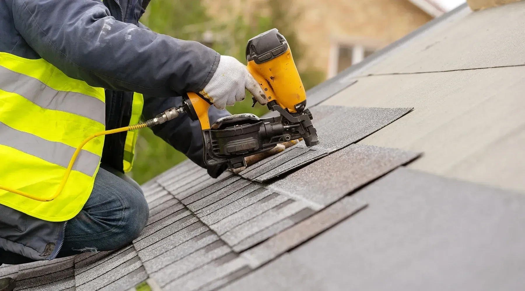 Worker cutting roof shingles with a power saw while kneeling on a sloped roof, wearing a high-visibility vest.