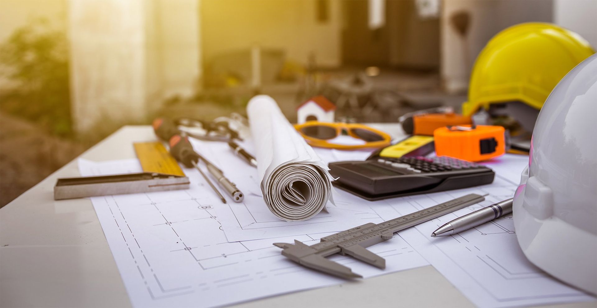 Construction plans and tools on a desk with a yellow hard hat and rolled blueprints