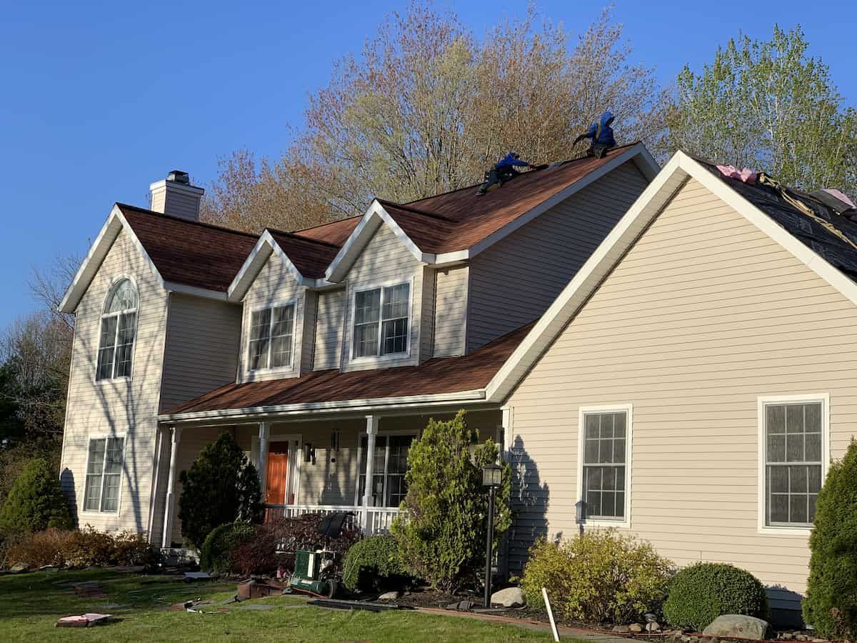 Suburban beige house with a red roof and front porch, surrounded by trees and shrubs.