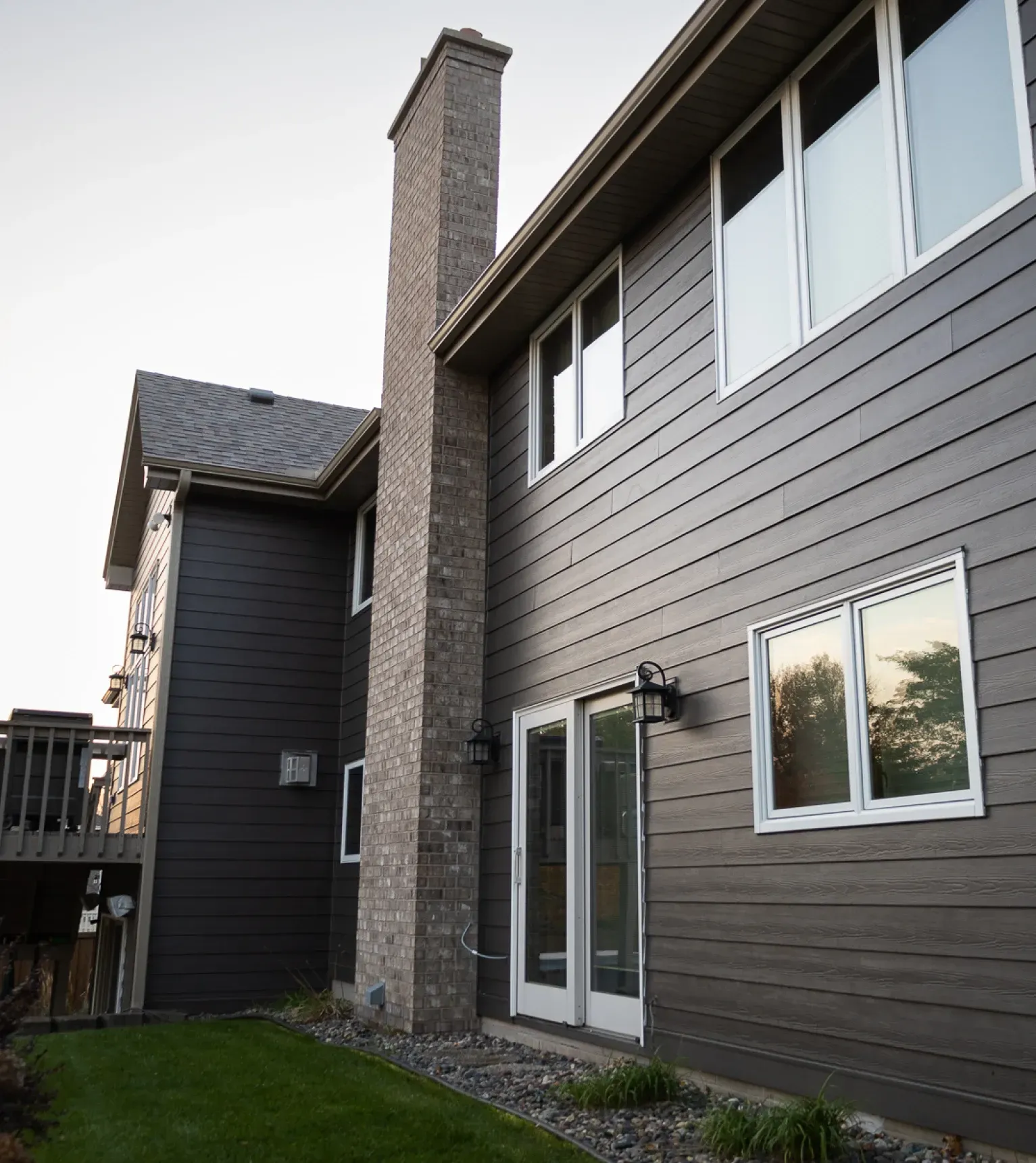Back view of a two-story dark gray house with brick chimney, windows, patio doors, and a small lawn