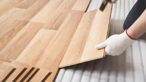 Gloved hands installing light wood laminate flooring over a corrugated underlayment