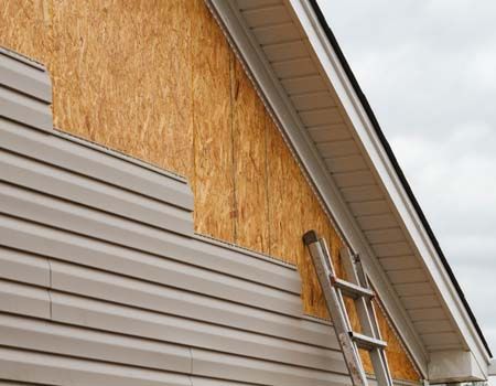 House exterior with ladder leaning against partially covered plywood wall and gray siding under the roofline