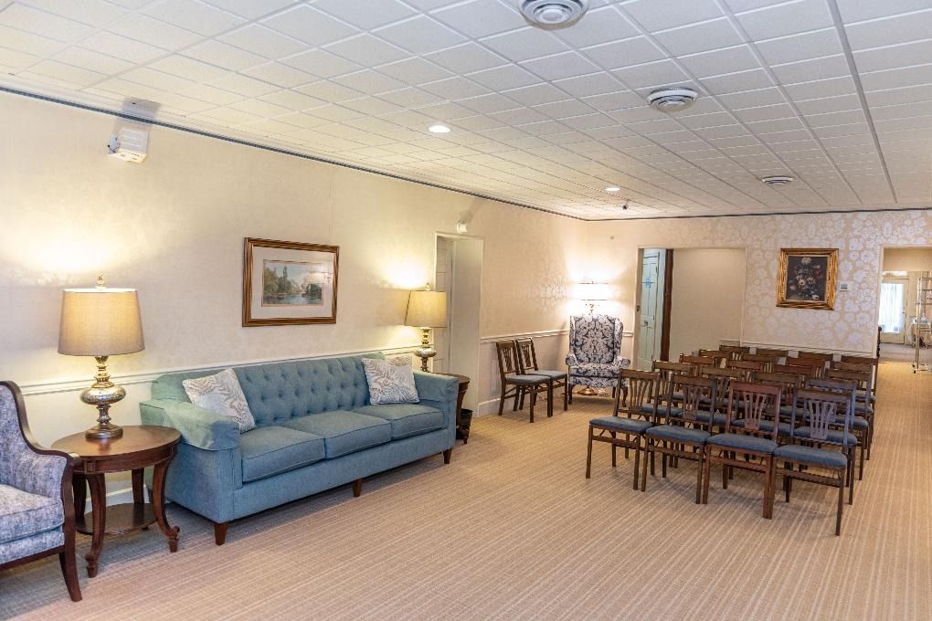 Spacious lounge with teal sofa, armchairs, lamps, and rows of wooden chairs on a beige carpet