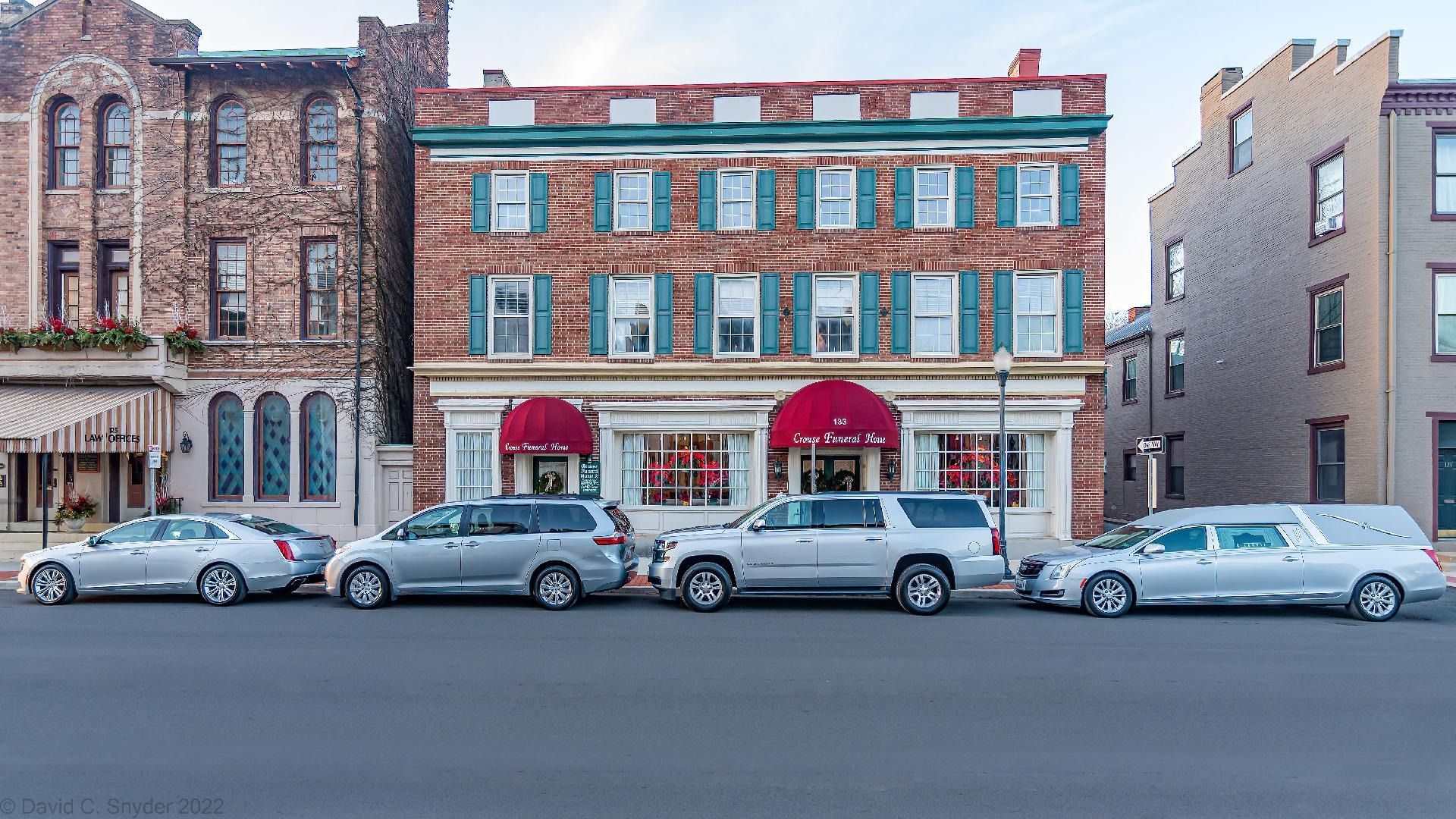 Historic storefronts with parked cars along a city street, centered on a brick building with red awnings.