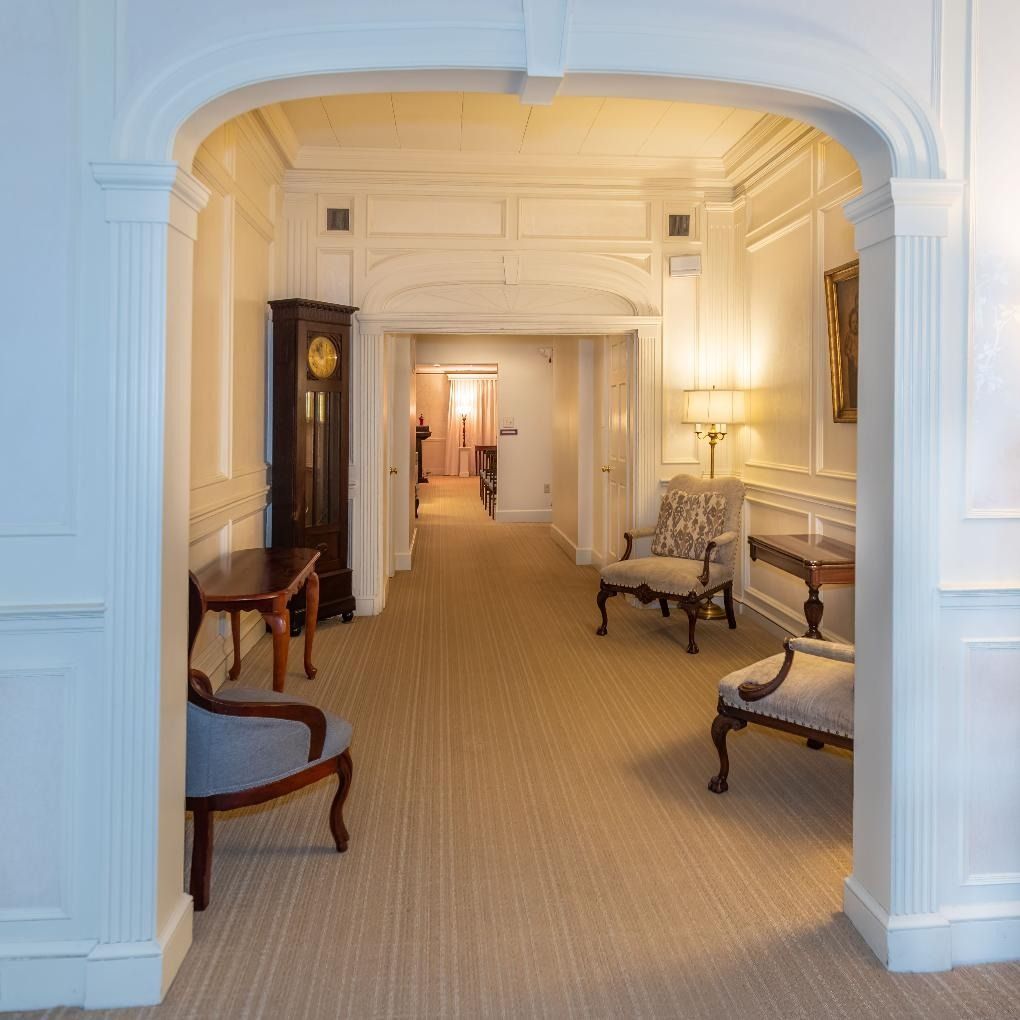 Elegant hotel corridor with white arches, carpet, and antique chairs leading to a softly lit hallway