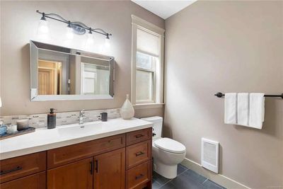 A bathroom with a wooden vanity, marble countertop, rectangular mirror, light fixture, window, and a toilet.