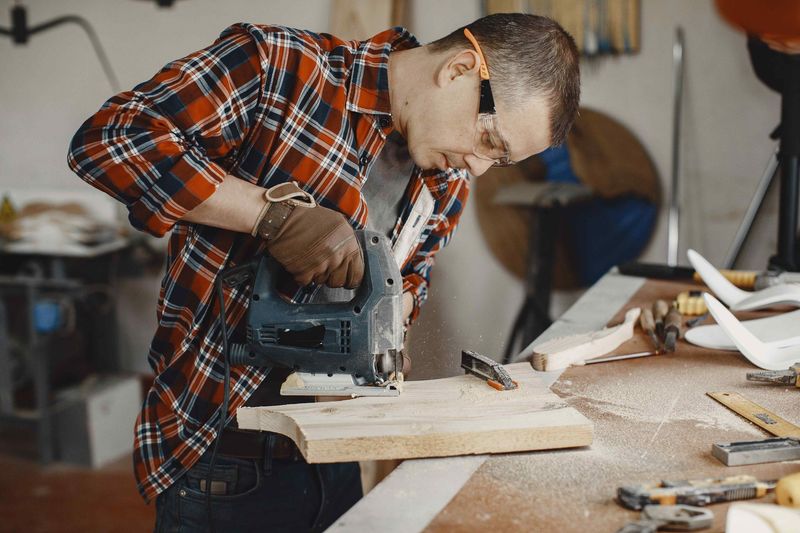 A person wearing a plaid shirt and safety glasses uses a jigsaw to cut a piece of wood in a workshop.