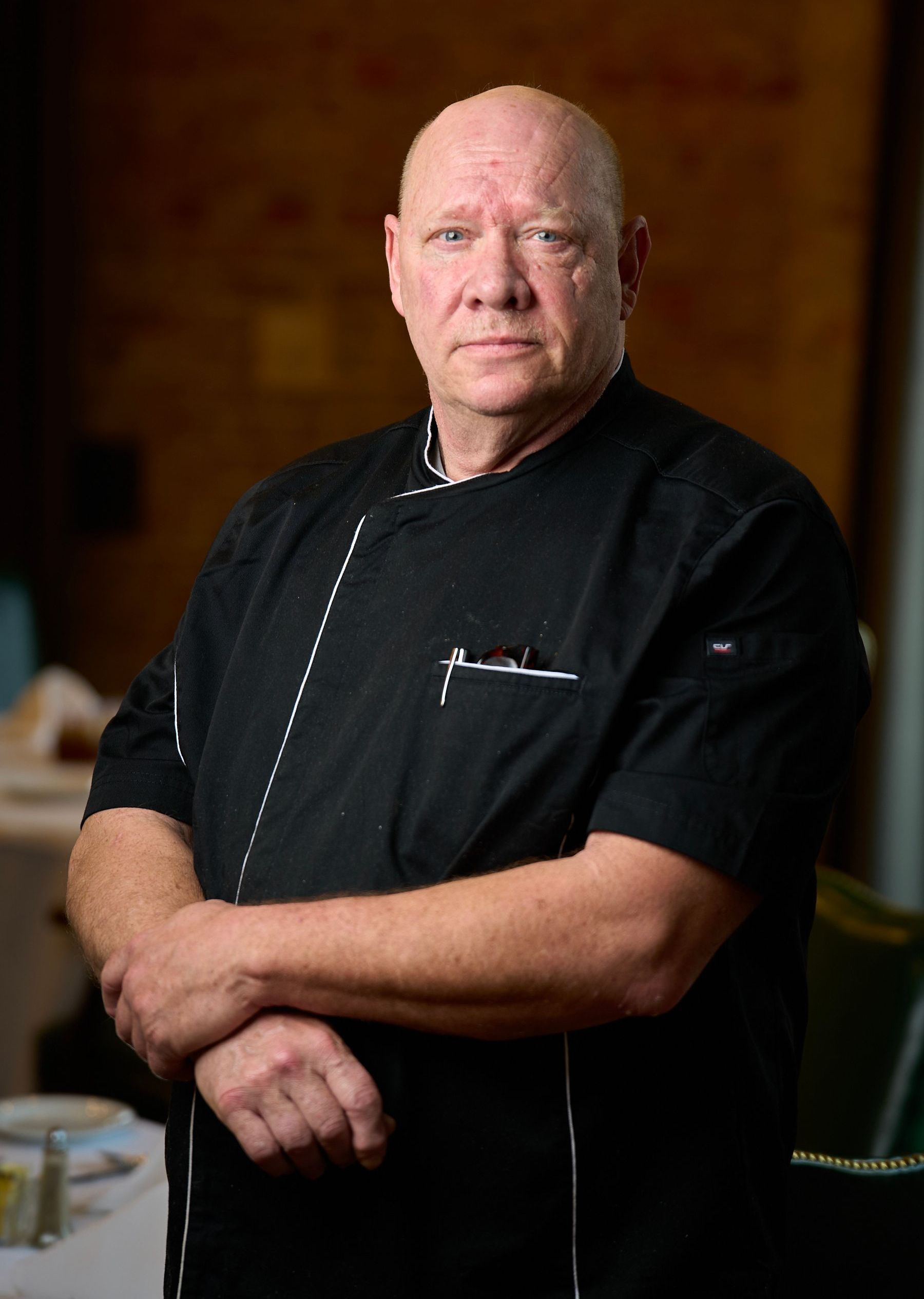 A bald man in a black chef 's jacket is standing with his hands folded.