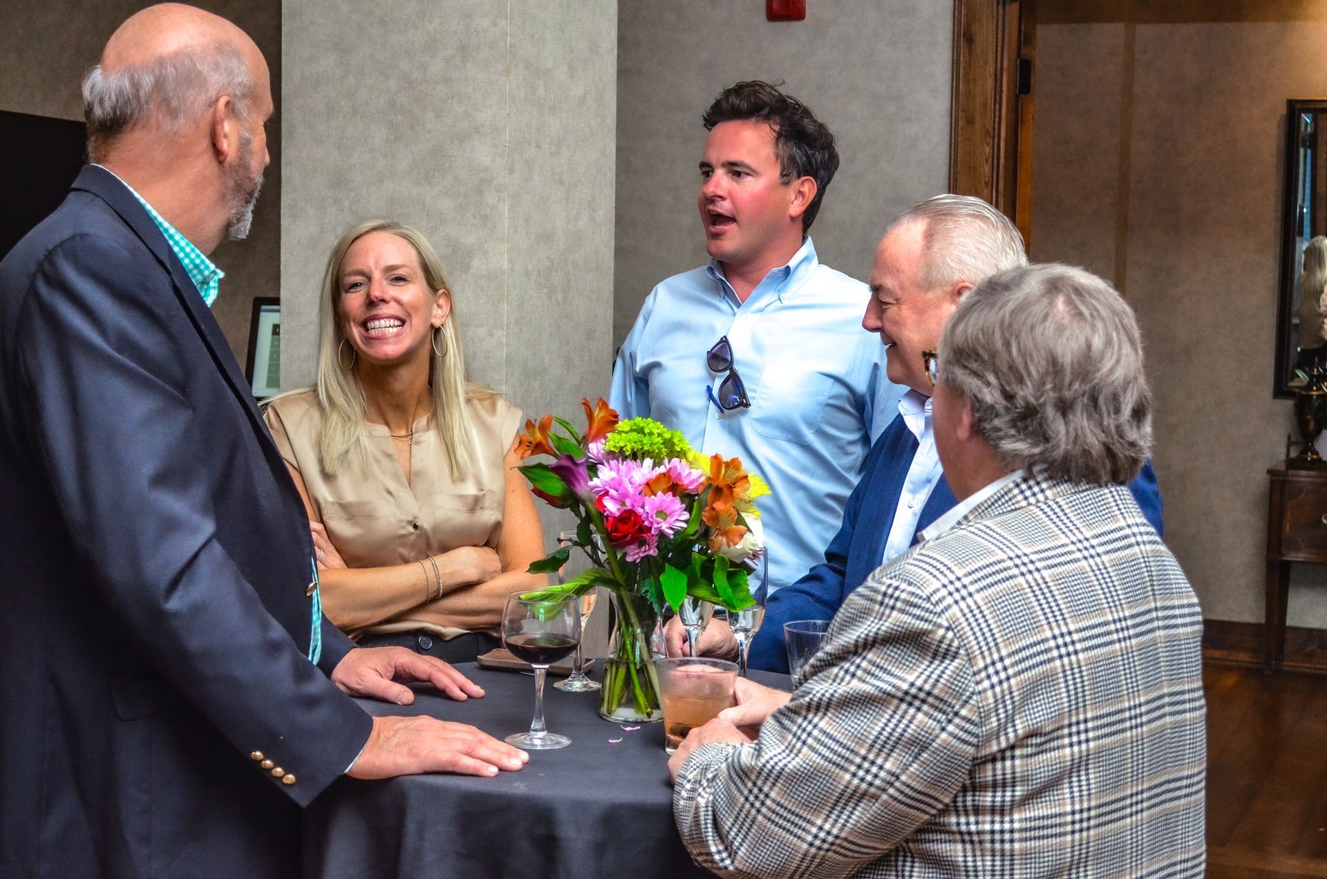 A group of people are standing around a table with a vase of flowers.