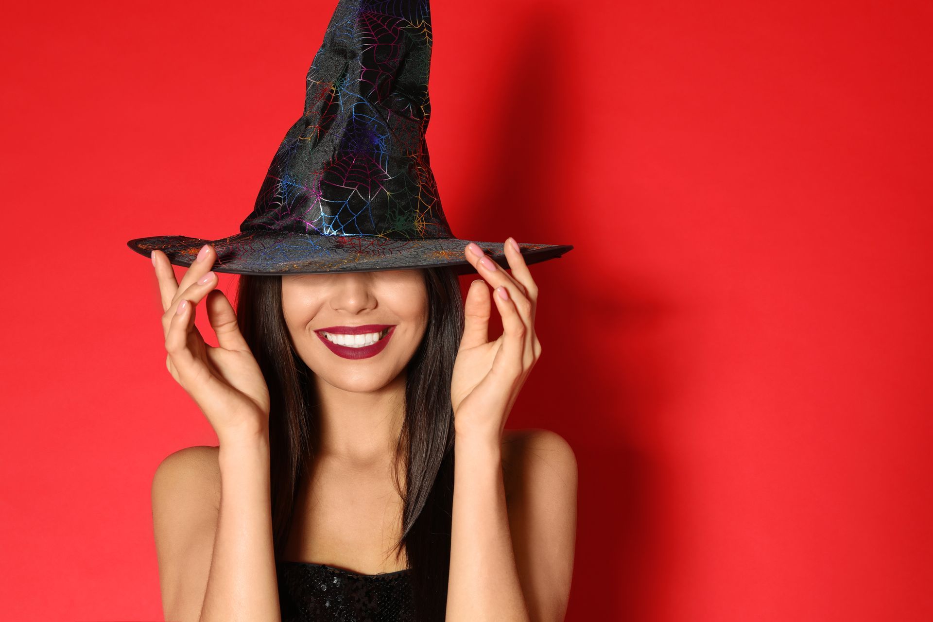 Woman wearing a shiny black witch hat,eyebrows hidden, smiling, red background.