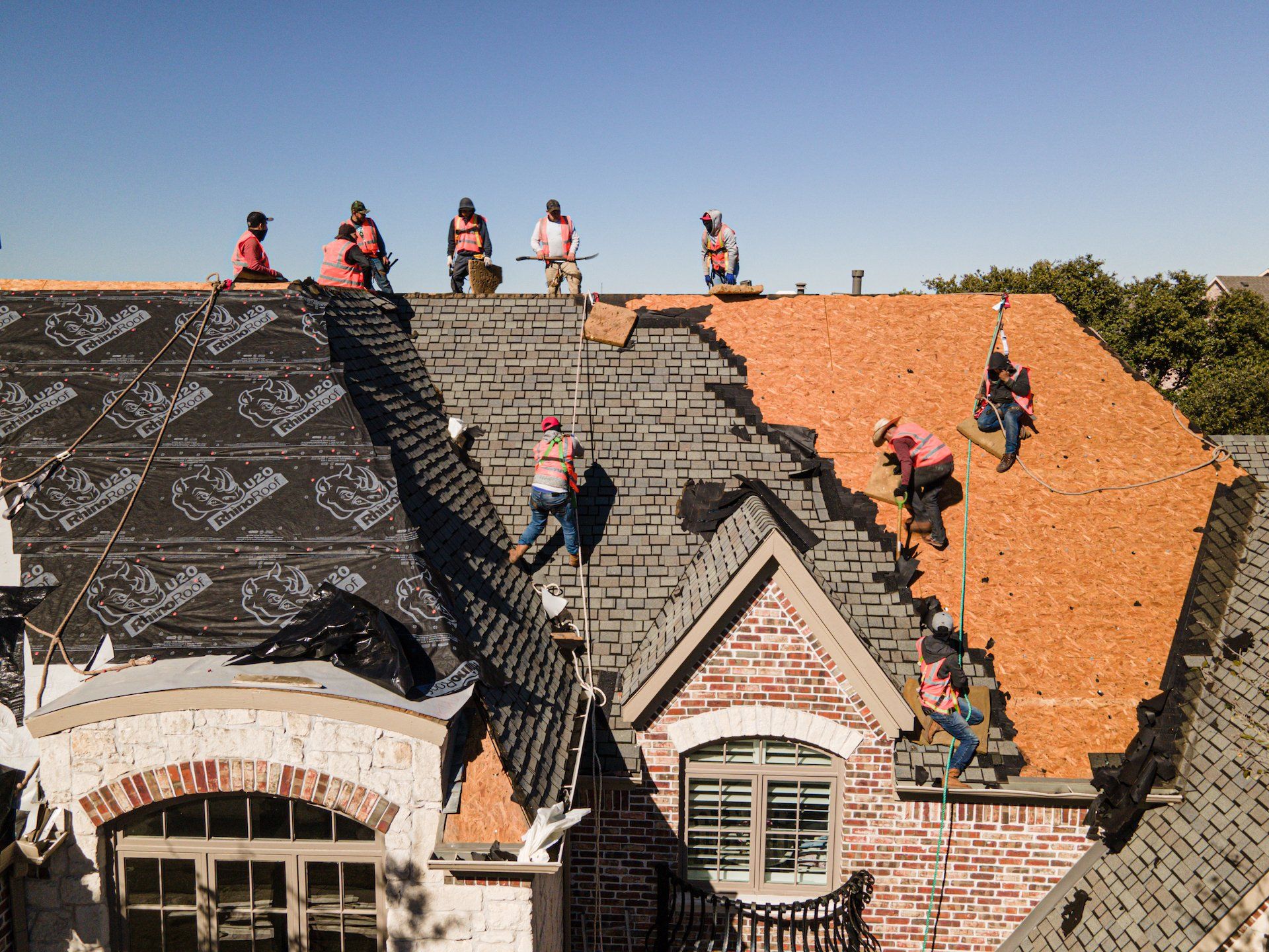 Workers installing dark and orange roof shingles on a house, with others on the roof ridge.