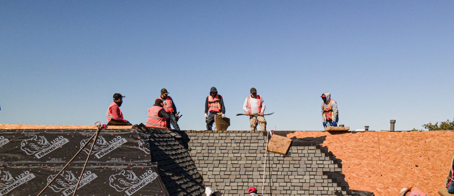 Roofers working on a roof under a blue sky, some wearing orange vests.
