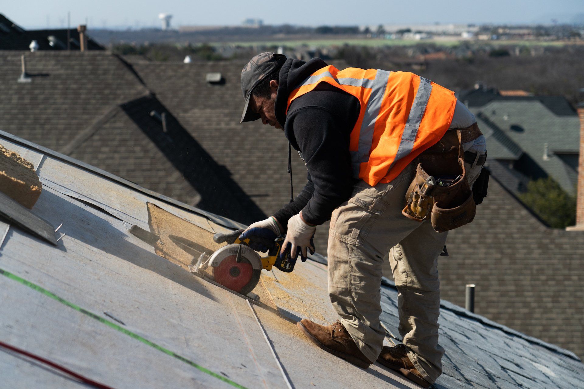 Worker in orange safety vest cutting roofing shingles with a circular saw on a sloped roof