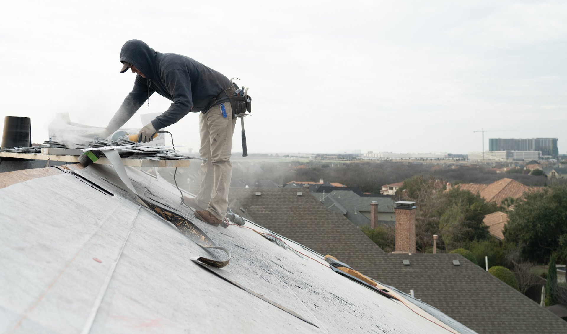 Roofer in a hooded sweatshirt cuts roofing material with a saw on a residential roof.