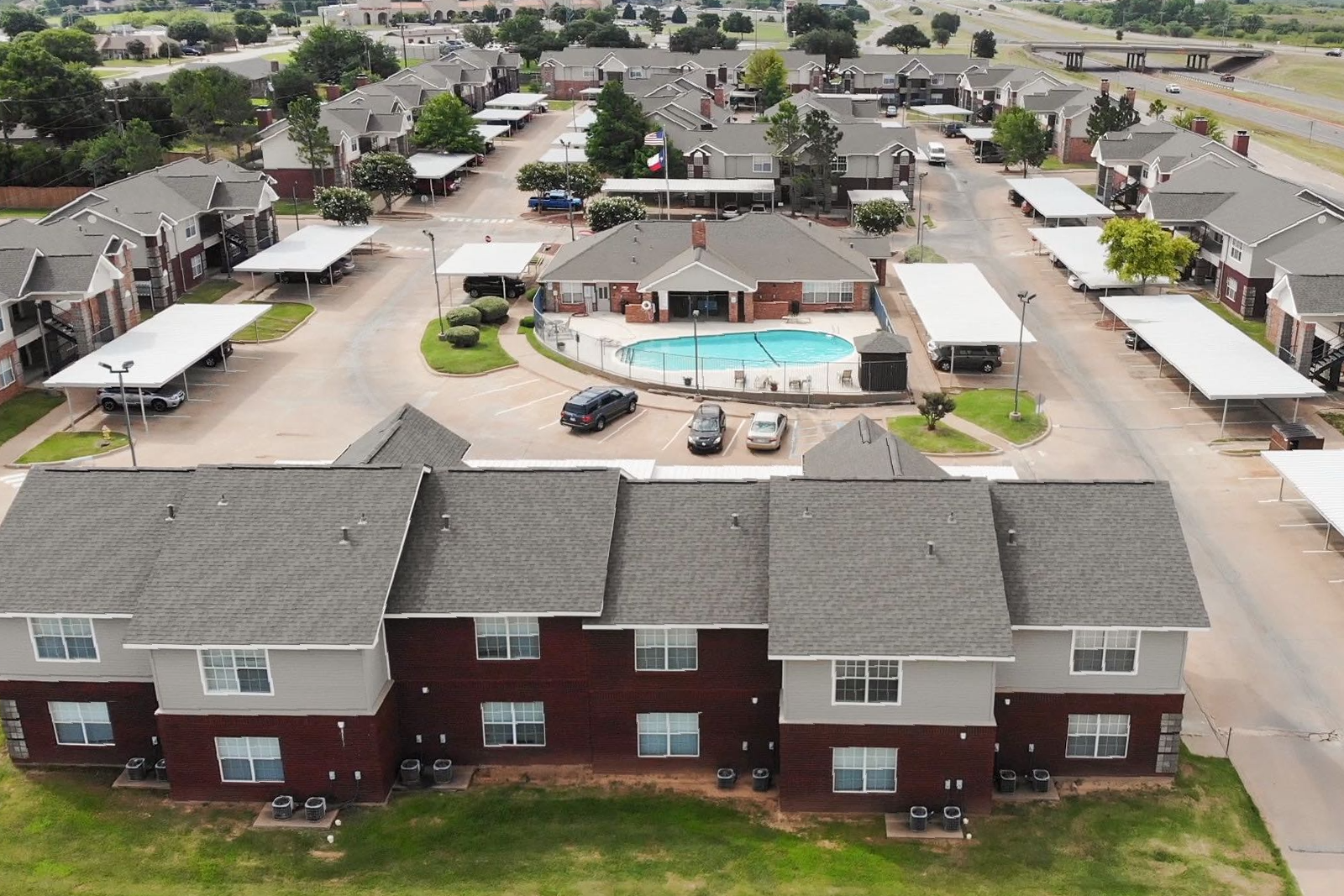 Aerial view of an apartment complex featuring residential buildings, a central swimming pool, and covered parking lots.