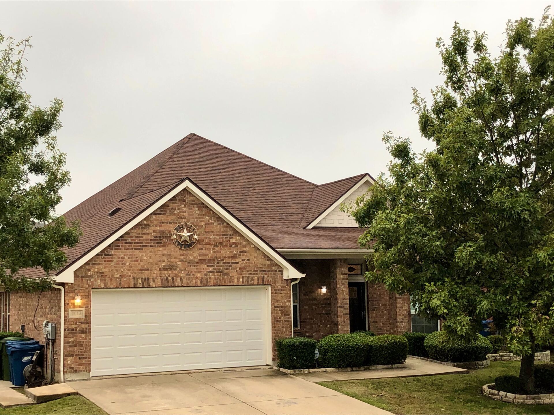 A single-story brick house with a brown shingled roof, a two-car garage, and green landscaping under a cloudy sky.