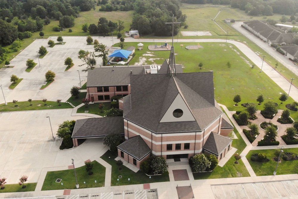 Aerial view of a large, octagonal-roofed church with a cross on top, surrounded by paved parking lots and green grounds.