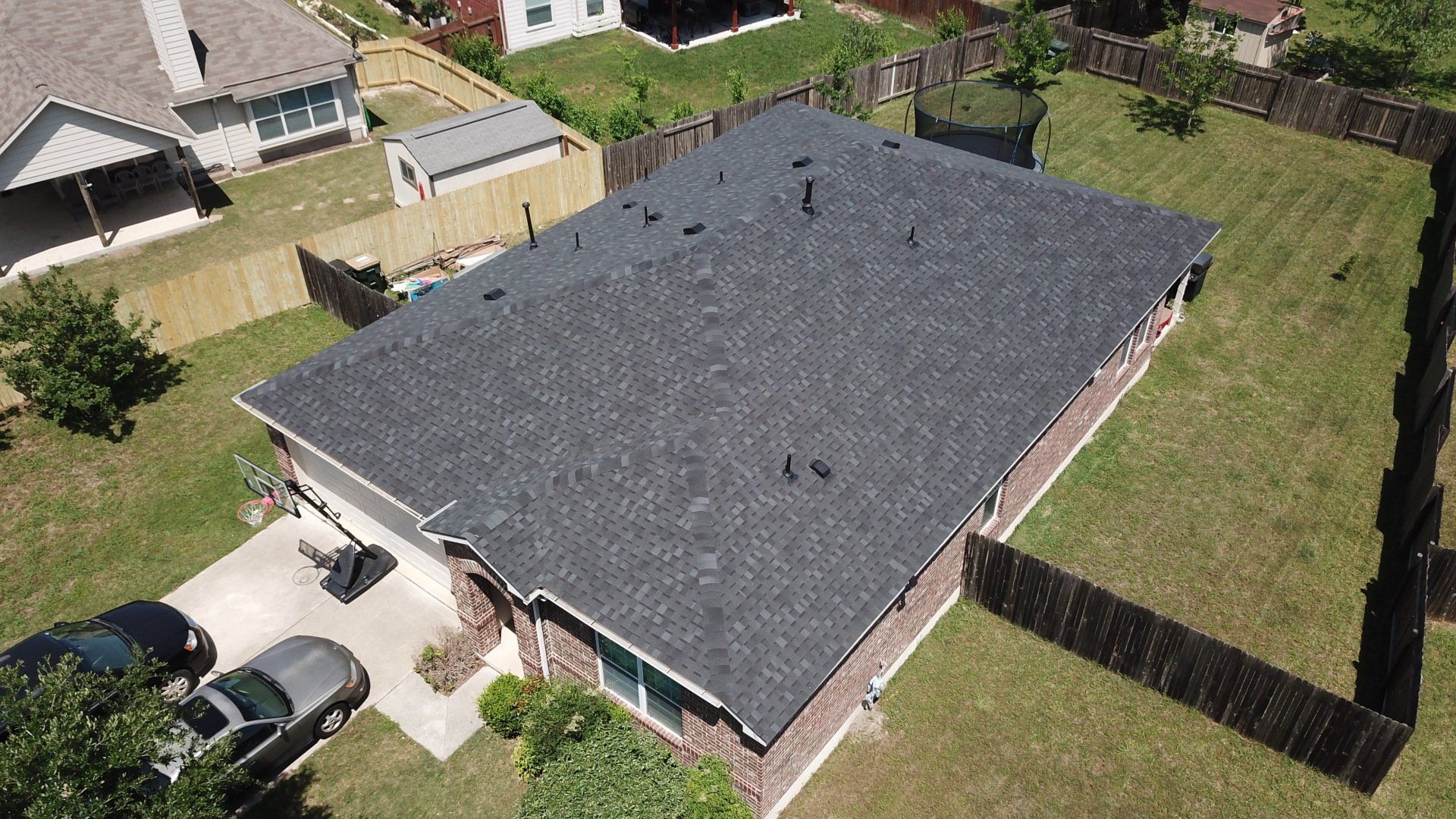 Aerial view of a single-story brick house with a dark gray roof and fenced backyard in a suburban neighborhood