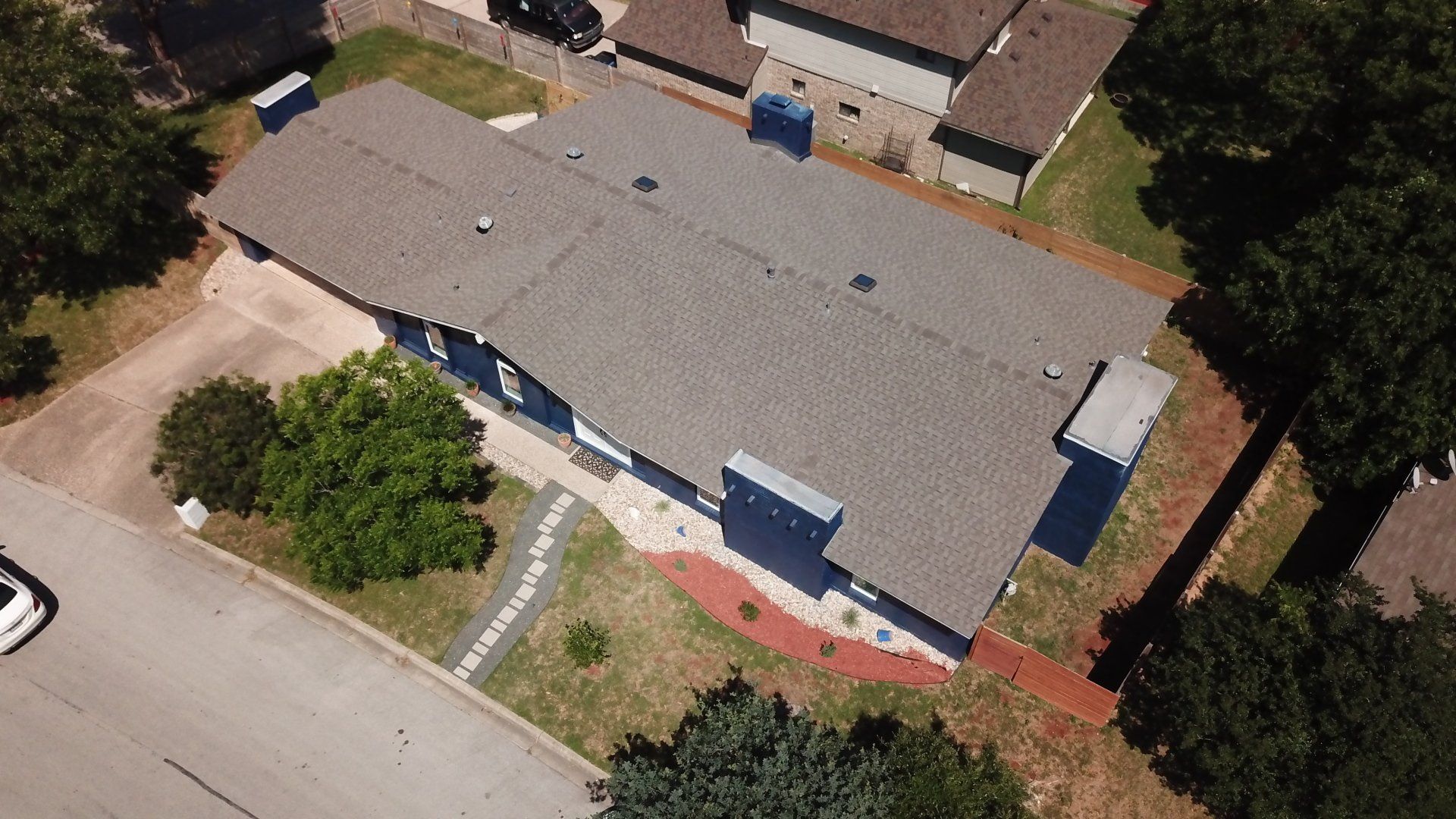 Aerial view of a single-story house with a gray roof and chimneys, surrounded by trees and a driveway.