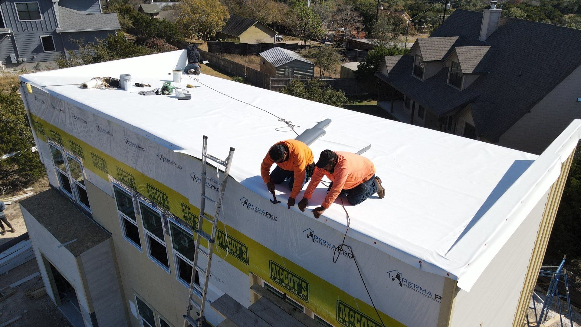 Two workers in orange safety gear install roofing on a white building roof.