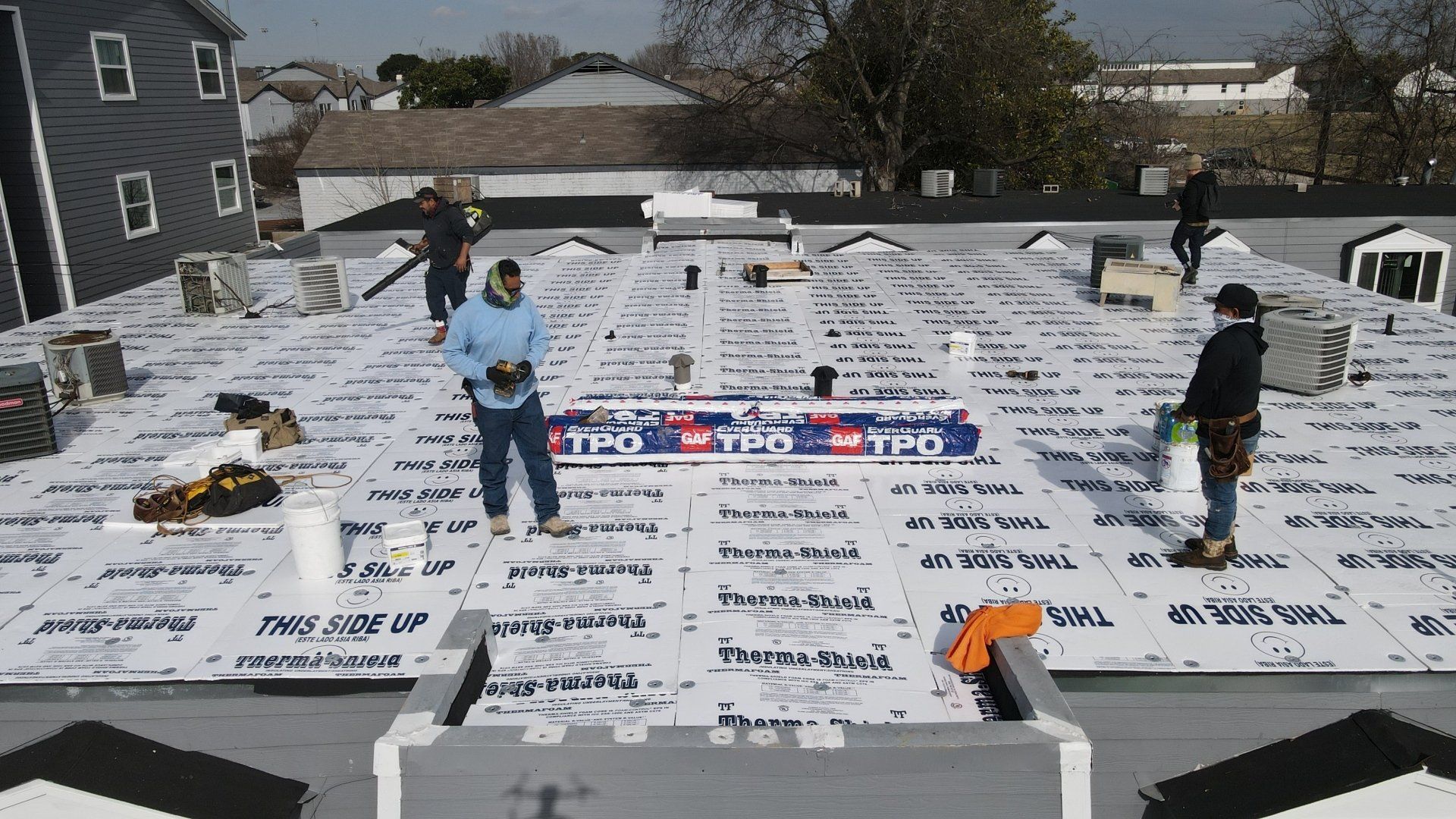 Workers installing roofing material on a flat roof. Several people are present; white material covers much of the surface.