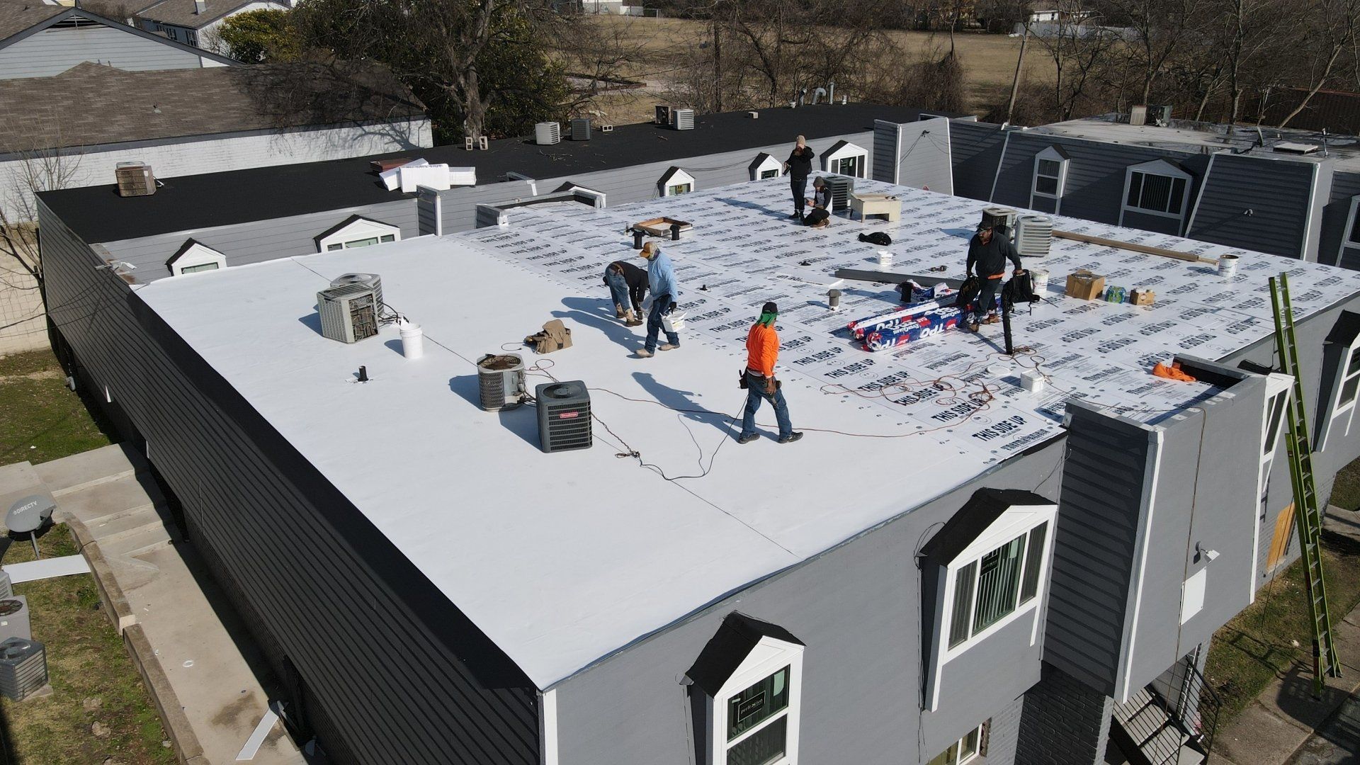 Workers installing roofing on a flat-roofed building. Gray building with multiple skylights and windows.