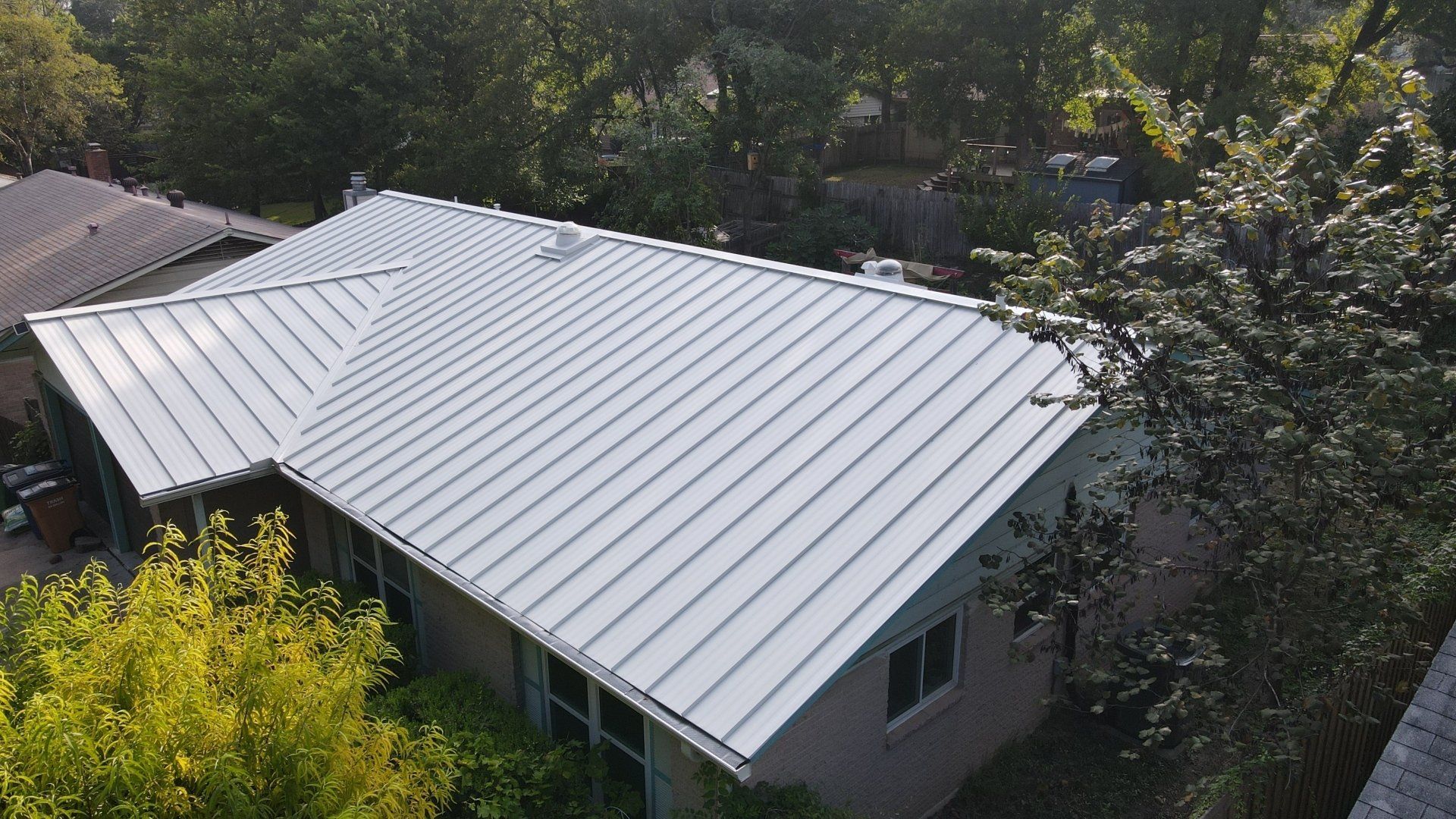 Silver metal roof on a house, viewed from above, surrounded by green trees.
